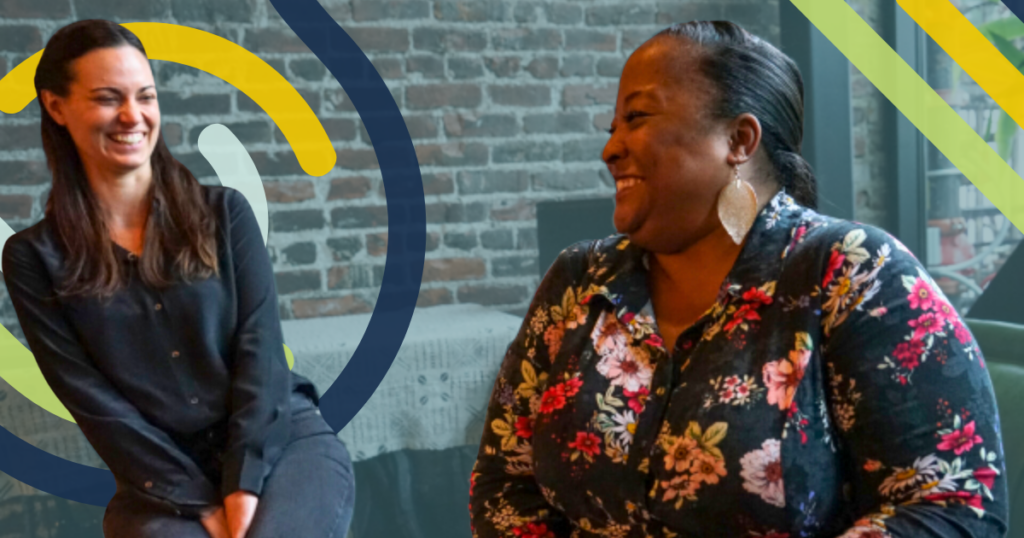 Two women sit next to each other in front of a semi-transparent background of a brick wall and share smiles and laughter.