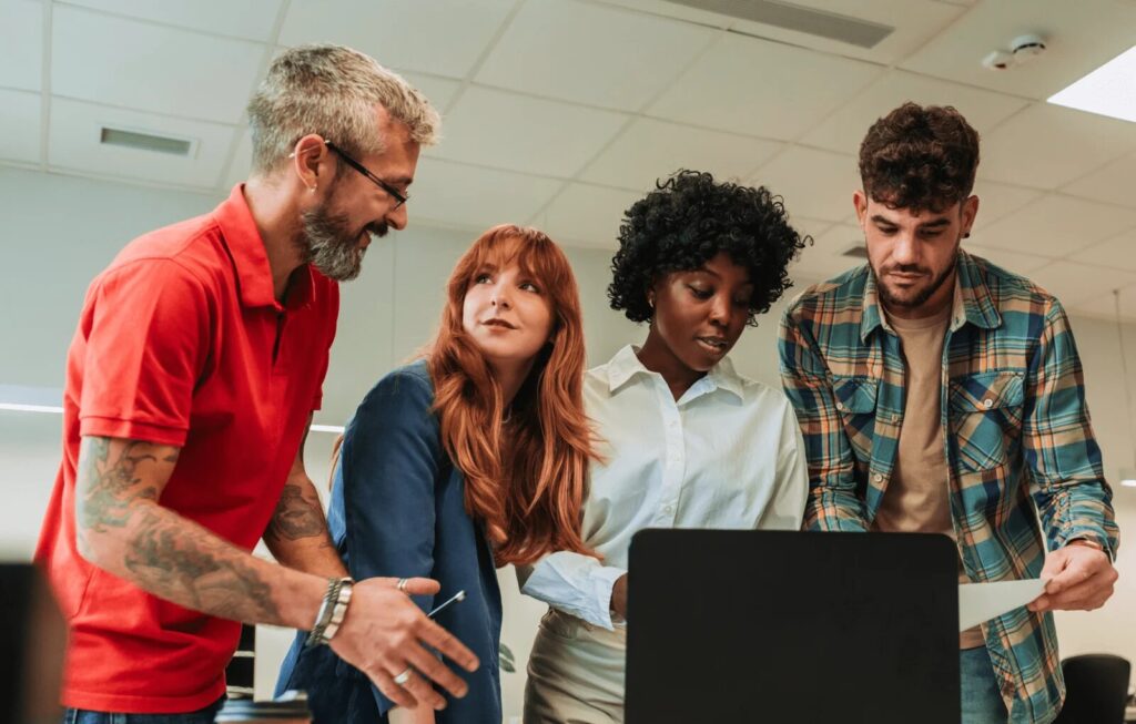 Two women and two men stand in front of a laptop computer and discuss what's on the screen.