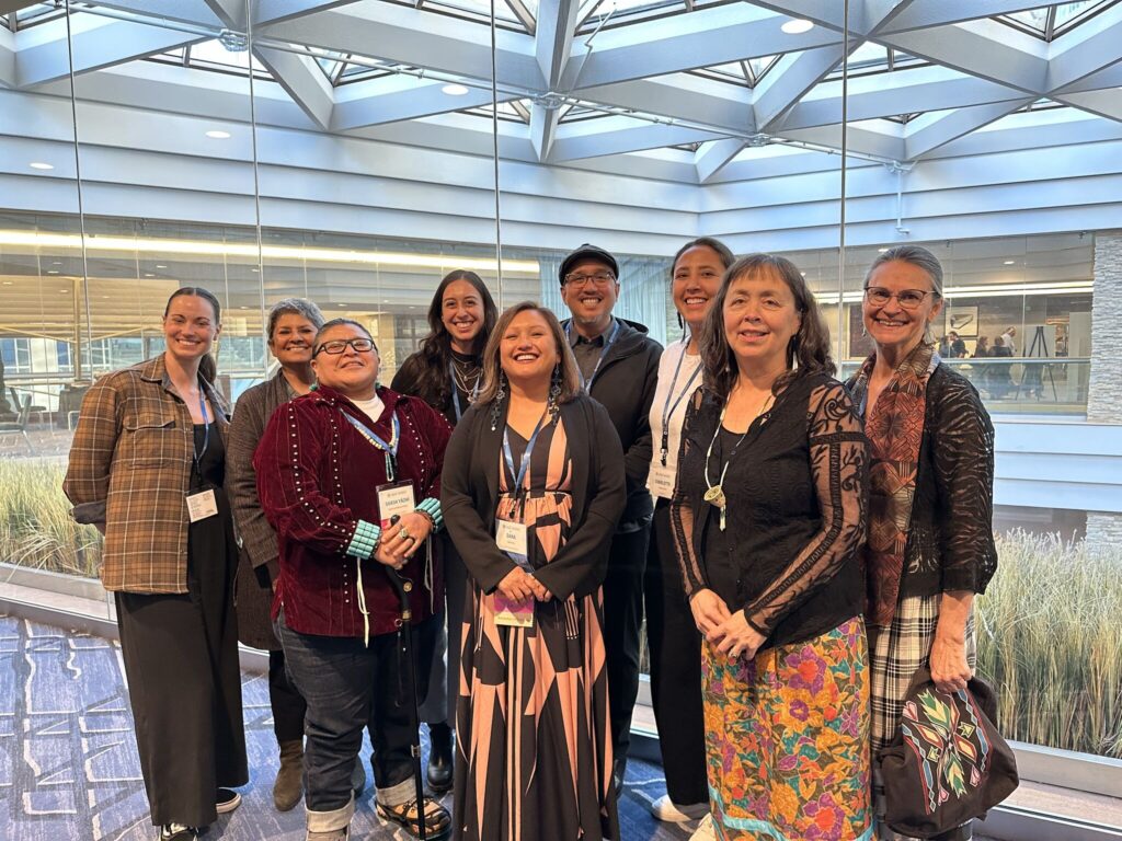 A group of nine people stand in front of a glass atrium with the sunlight beaming in overhead.