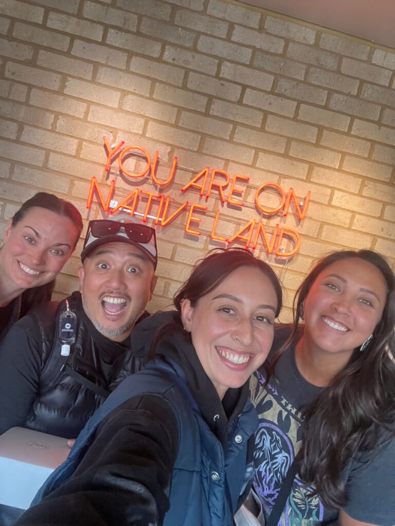 Four people stand in front of a brick wall with a neon sign that says you are on Native land.