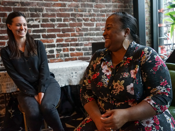 Two women sit and smile at each other in front of a brick wall in the background.